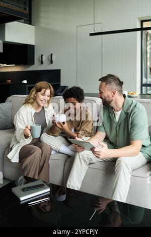 Group of friends having relaxed conversation while sitting on couch in modern living room and enjoying coffee. Three friends sharing happy moment while smiling and holding cups Stock Photo