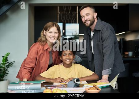 Family smiling together at table with books and snacks, with supportive parents standing beside cheerful child with loving expressions in cozy home setting Stock Photo