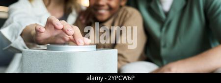 Family members gathered around smart speaker, using modern technology to enhance home experience. Focus on hand and fingers touching device controls Stock Photo