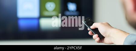 Hand gripping TV remote control in front of blurred television screen displaying various icons and applications Stock Photo