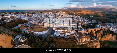 Aerial image of Ronda, Spain, showcasing Plaza de Toros, El Tajo gorge, and Puente Nuevo bridge at sunset, with white buildings and distant mountains. Stock Photo