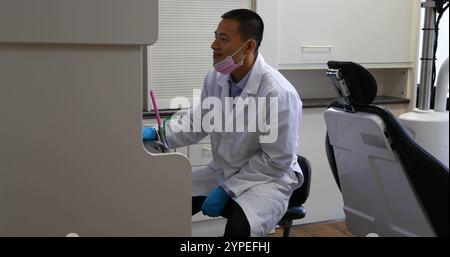Side view of smiling male Asian dentist wearing lab coat using desktop PC in clinic Stock Photo