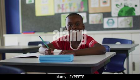 Portrait of happy african american schoolboy sitting at classroom, making notes, looking at camera Stock Photo