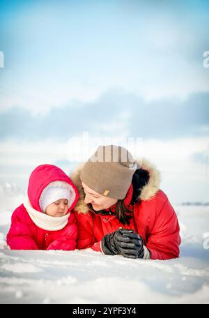 Happy mother and baby daughter wearing warm winter clothes lying on snow and playing outdoors on beautiful winter day Stock Photo