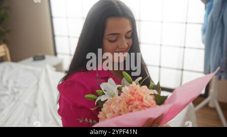 Young woman in a bedroom holding flowers, wearing a pink shirt, captured in an indoor home setting with a soft smile on her face Stock Photo