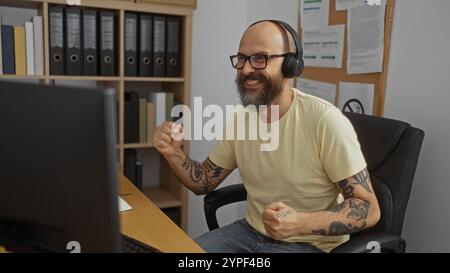 Hispanic man with tattoos and beard celebrating at his desk in an office workplace, wearing headphones and glasses, with shelves of binders in the bac Stock Photo