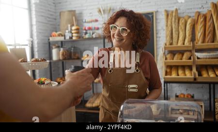Woman baker smiling and serving coffee to customer in cozy indoor bakery with freshly baked bread in background showcasing the friendly interaction be Stock Photo