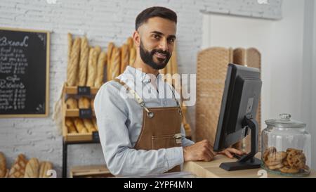 Handsome young hispanic man in apron working behind the counter at a bakery shop with bread in the background and a jar of cookies on the counter Stock Photo