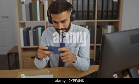 Young man with beard using a smartphone while wearing headphones in a modern office, surrounded by shelves and folders, appearing focused and engaged Stock Photo