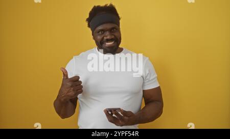 A handsome young african american man gives a thumbs-up while holding a smartphone, standing against a vibrant yellow background. Stock Photo