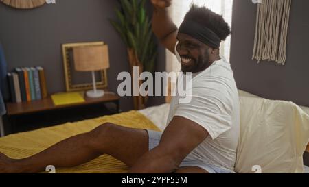 A young handsome african american man with a beard, sitting up in bed in a cozy bedroom, stretches and smiles, surrounded by a serene indoor setting. Stock Photo