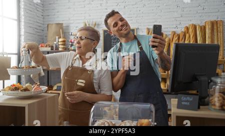 Man and woman bakers taking selfie in bakery while arranging pastries on tray indoors. Stock Photo