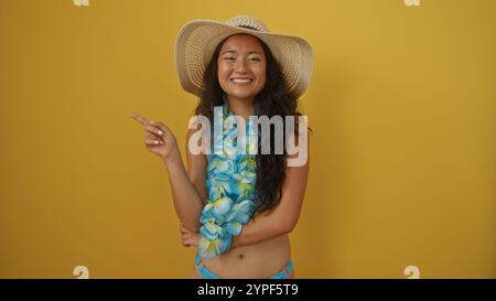 Beautiful chinese woman wearing beachwear and a hat, smiling and pointing, isolated against a vibrant yellow background Stock Photo