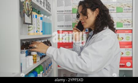 A young hispanic woman pharmacist, wearing a white coat, is talking on the phone while arranging shelves in a well-stocked pharmacy or drugstore inter Stock Photo