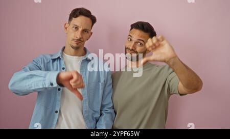 Gay couple standing together making thumbs down gestures against a pink background, embracing their lgbtq representation with casual attire and facial Stock Photo
