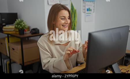 Attractive young blonde caucasian woman working at her desk in an office setting, smiling as she interacts with her computer in a modern workplace env Stock Photo