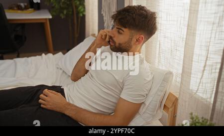 Young man with beard relaxing on bed in bedroom talking on phone in cozy home interior Stock Photo