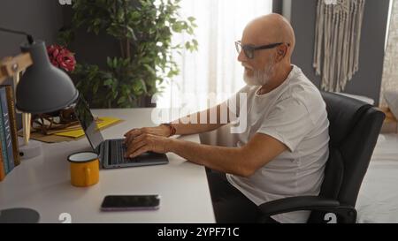 Man typing on laptop at desk in bedroom with grey walls, indoor plants, books, lamp, and cup, wearing glasses and white t-shirt, showing a mature, mid Stock Photo