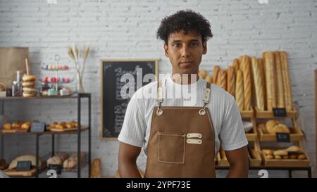 Hispanic man working in an indoor bakery, surrounded by fresh bread and pastries, wearing a white shirt and brown apron, with a neat interior showcasi Stock Photo