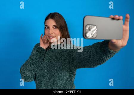 Young woman taking a selfie with smartphone on blue background, touching her hair and smiling Stock Photo