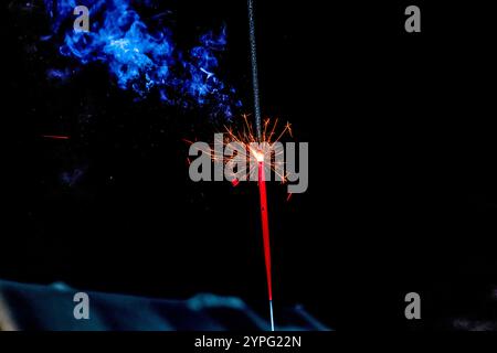 Lit sparkler glowing against black night background with vibrant blue smoke and scattered sparks. Sweden. Stock Photo