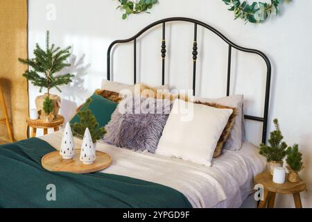 Cozy bedroom setup with festive Christmas decor. Bed with forged headboard, green blanket, plush pillows and small decorative trees on wooden tray.  F Stock Photo