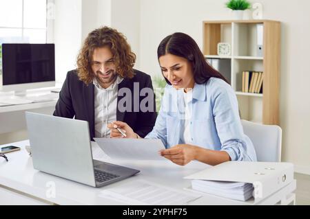 Two business accountants sitting at an office desk together, doing paperwork and using a laptop Stock Photo