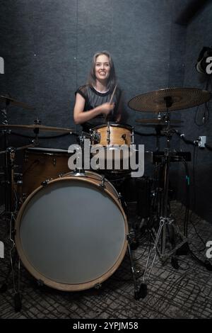 Smiling female drummer performing on drum set in professional soundproof studio. Vibrant scene emphasizes energy, creativity, and joy of live music creation, vertical image Stock Photo