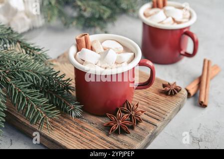 Two red enamel mugs with hot chocolate, marshmallows and cinnamon sticks on a wooden cutting board with fir branches on the background Stock Photo