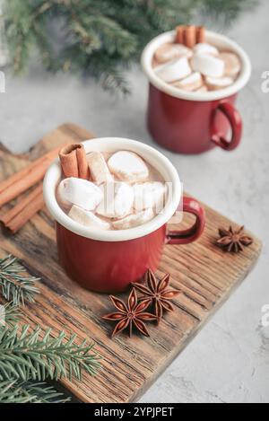 Two red enamel mugs with hot chocolate, marshmallows and cinnamon sticks on a wooden cutting board with fir branches on the background Stock Photo