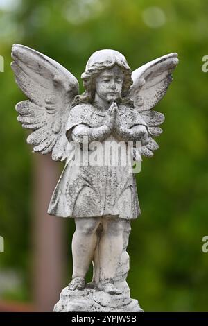 Portrait of predominantly white stone sculpture or statue of a cherub, or an angel, with its hands clasped together in prayer, in a cemetery Stock Photo