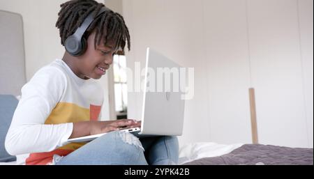Happy african american boy wearing headphones sitting on bed using laptop at home, slow motion Stock Photo