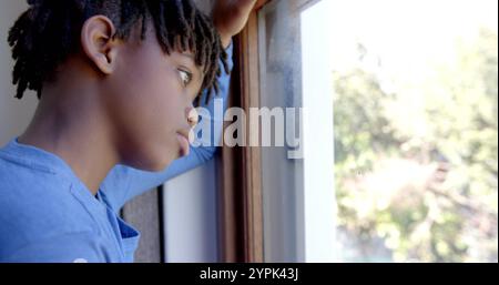 Thoughtful african american boy looking through window at home, slow motion Stock Photo
