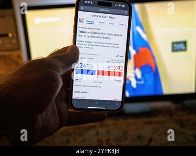 Paris, France - Nov 13, 2024: A hand holds a smartphone displaying live US presidential election results on the Financial Times website, showing Trump leading over Harris with 292 to 224 electoral votes. A TV with election visuals is in the background. Stock Photo