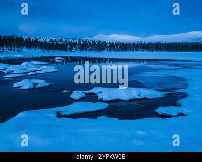 A tranquil winter evening shows dark waters while snow-covered mountains rise in the background. The blue dusk light enhances the beauty of this remot Stock Photo