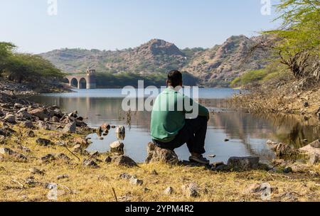 Young Man Sitting on Stone by Pristine Lake at Morning Stock Photo