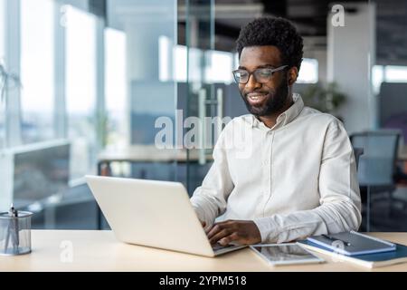 African American man businessman engaged in work on laptop in bright office. Casual shirt and glasses add charm to professional setting. Light filled space enhances productivity and focus. Stock Photo