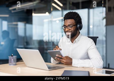 African American businessman engaged in online learning using laptop and notebook. Wearing headphones, he is focused and smiling in modern office environment. Stock Photo