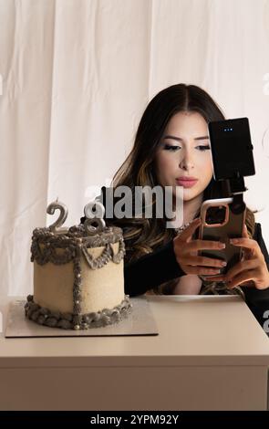 A woman is taking a picture of a cake with the number 28 on it. She is holding a cell phone and she is focused on capturing the cake's details Stock Photo