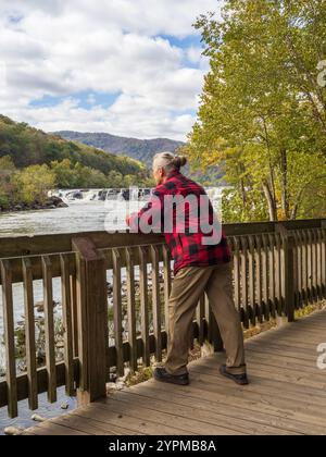 A man in a red lumberjack jacket and hair bun stands in awe of Sandstone Falls, surrounded by the vibrant autumn colors of New River Gorge National Pa Stock Photo