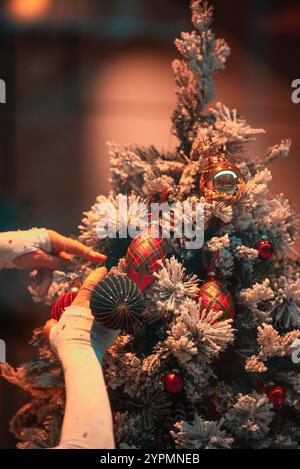 Warm orange-toned Christmas scene with festive lights and ornaments on a beautifully decorated tree. A lady’s hands are gently adding decorations Stock Photo