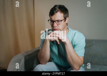 Young man wearing glasses is sitting thoughtfully on a sofa, his hands clasped under his chin, lost in deep thought, reflecting on life's complexities Stock Photo
