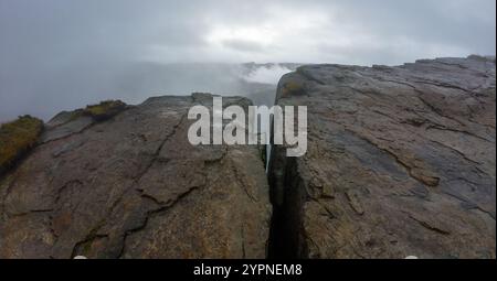 A dramatic landscape featuring a large rock formation with a deep crevice, surrounded by mist and fog, creating a mysterious and moody atmosphere. Stock Photo