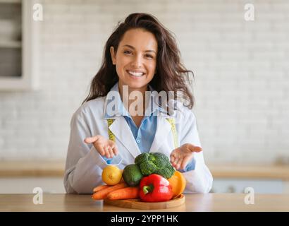 Portrait of cheerful dietitian in lab coat pointing at fresh fruits and vegetables on table, smiling at camera at clinic Stock Photo
