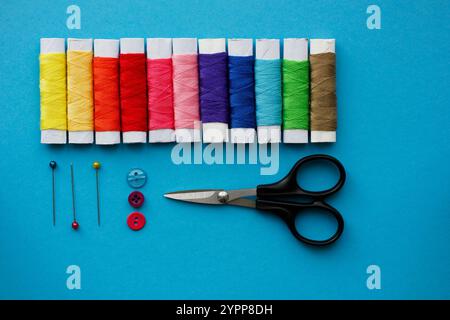 Composition of sewing accessories on blue background. Multicolored sewing threads, scissors, pins and buttons. Top view, simple flat lay. Stock Photo