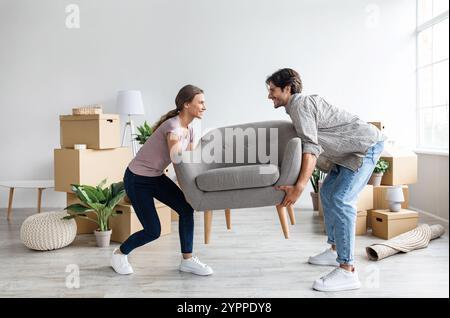Smiling caucasian millennial wife and husband carry armchair in room with cardboard boxes Stock Photo