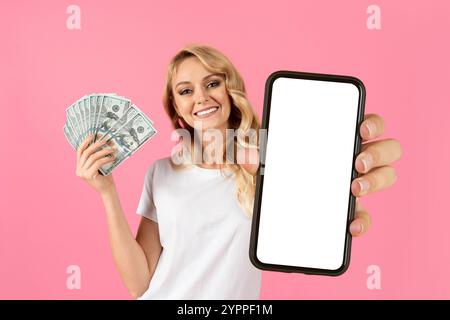 Young woman smiles while holding cash and a blank phone against pink backdrop Stock Photo
