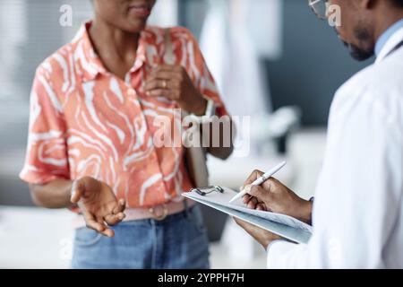 Side view cropped shot of African American doctor holding clipboard and taking notes while listening to female patient in clinic, copy space Stock Photo