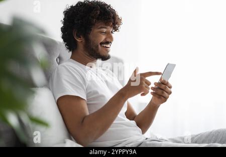 Positive dark-skinned guy scrolling in bed, using smartphone Stock Photo