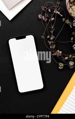 Modern smartphone on black table background with fragile houseplant decoration, yellow and gray notepads - stylish workspace setup. Top view. Stock Photo